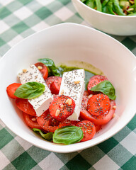 Caprese salad with strawberry, mozzarells, basil and balsamic sauce served in a white bowl over green plaid background.