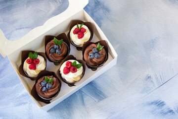 Cupcakes  with cream cheese and chocolate cupcakes with fresh blueberries and raspberries in  white paper box, blue background. Berry cupcakes in delivery box. Sweet food delivery