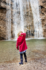 Kind steht vor Wasserfall. Mädchen spielt mit Steinen in der Natur vor Wasserfall. Child stands in front of a waterfall. Girl plays with stones in nature in front of waterfall.