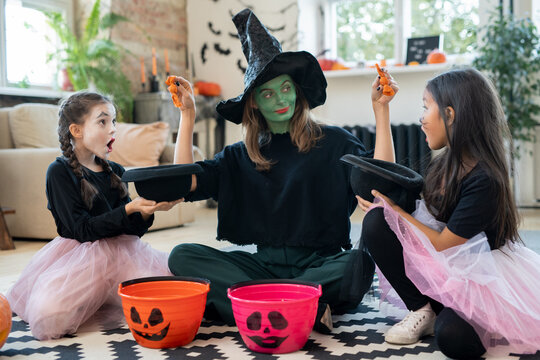 Young female in witch attire putting treats in hats of two adorable girls while sitting on the floor