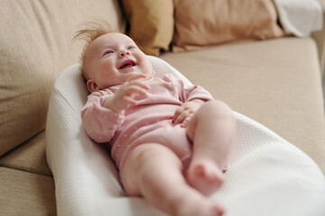 Happy cute baby girl in pink loose jacket laughing while lying in small soft bed on sofa