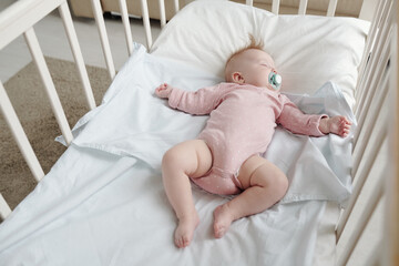 Serene barefoot baby girl in pink clothes lying on white linens in cradle