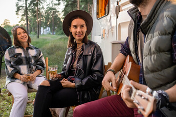 Two happy girl with drinks listening to young man playing guitar by house on wheels in the countryside