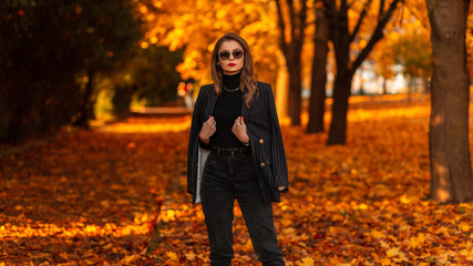 Cool business woman model with stylish sunglasses in fashionable black suit clothes with fashion blazer walks in the autumn park with orange foliage