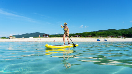 Woman stand up on paddle board in sea. Big yellow board in turquoise water. - obrazy, fototapety, plakaty