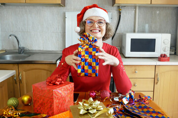 Woman preparing gifts for Christmas and New Year at home in kitchen, smiling woman in santa helper...