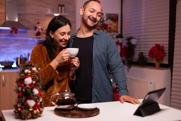 Happy couple celebrating christmas holiday with remote friends during online videocall meeting on tablet. Smiling family enjoying spending winter season time together in xmas decorated kitchen