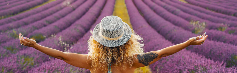 Rear view of beautiful woman standing on lavender flower field with arms outstretched. Woman in hat...
