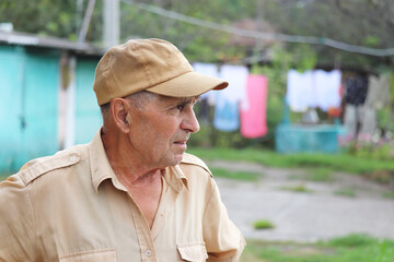 Elderly man standing in rural yard on background of well and hanged laundry. Concept of life in village, old age
