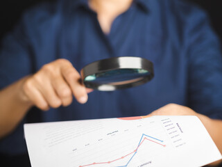 Businessman holding a magnifying glass looking at the report for analytics and statistic. Paper...