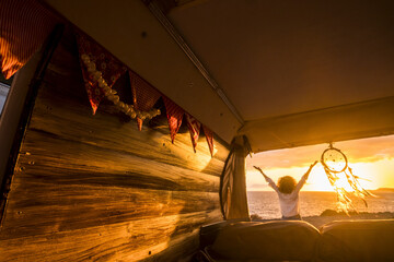 Rear view of woman with arms outstretched admiring scenic seascape during sunset next to dreamcatcher hanging from the camper van. Woman with curly hair enjoying her vacation at beach by dreamcatcher