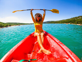 Obraz premium Happy caucasian woman holding oar sitting on red canoe in lake during sunny day. Beautiful female tourist holding oar with both arms raised having fun on a canoe ride during summer