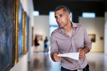 Adult man is visiting museum with information brochure and looking at the pictures in the gallery indoor.