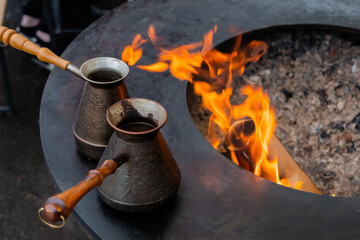 Preparing Turkish coffee with black cezve or ibrik - traditional coffee pot on outdoor round brazier with hot flame at street food festival - close up