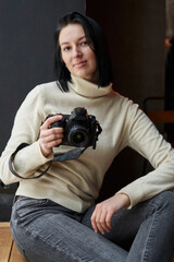 young beautiful woman in a light sweater sits by the wall and holds a camera in her hands