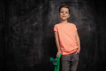 Schoolboy skateboarder with green skateboard on background of black chalk board with copy space. Stylish athletic boy ready to skateboarding. Childhood and active urban lifestyle.