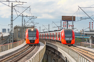Two electric passenger train drives at high speed among urban landscape.