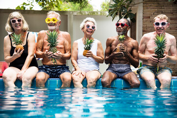 Group of diverse senior adults sitting at poolside holding pineapples together - Powered by Adobe