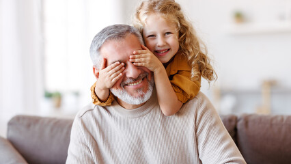 Cute little girl covering eyes with hands of her smiling grandfather while playing together at home