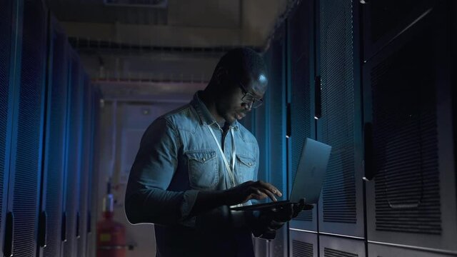 American engineer using laptop and typing while standing in dark room at data center spbas. Young African man holds computer in hand and types, examines racks of equipment or hardware and stands