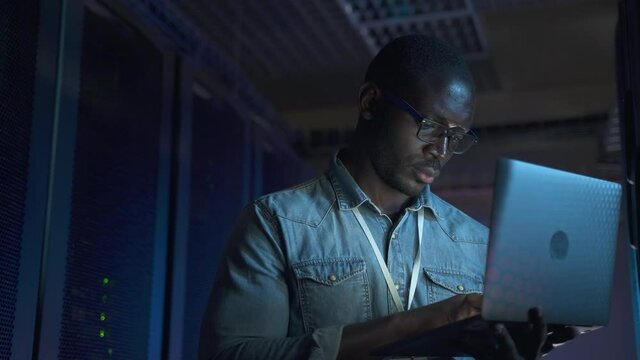 IT specialist using laptop and checking networks during working hours in render farm spbas. Close-up view of American African man holds computer in hand and does analytical job, inspects equipment or