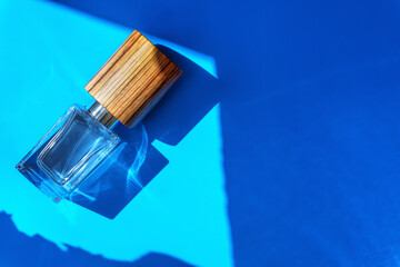 Luxury perfume glass bottle with a wooden cap on the light blue paper background. Rectangle cosmetic jar in sunlight 