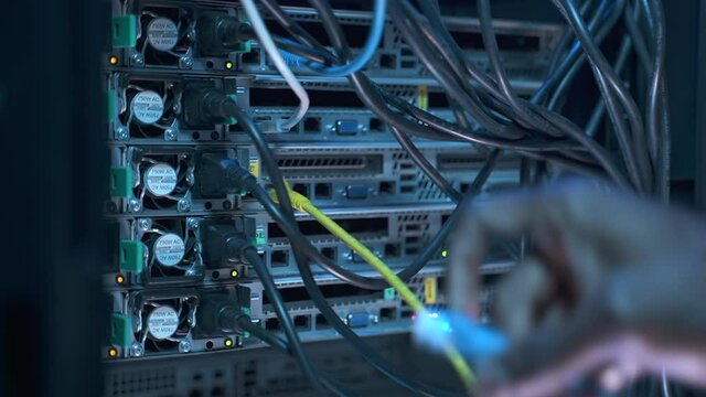 Closeup of man engineer working with cables and networks during dark night on render farm spbas. Male hand inserts cable into operating equipment or hardware during working hours in server room. Young