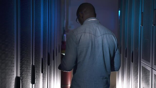 Engineer working with laptop and walking along interior during working day at date office spbas. African American man holds computer in hand and types, examining equipment racks and walks in dark room - Powered by Adobe