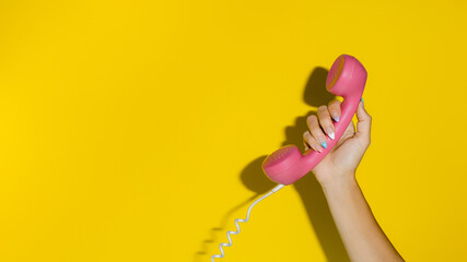 A woman's hand holding a pink vintage telephone speaker isolated on vibrant yellow background. Creative message or communication concept. Pop art visual trend and hard shadows with copy space.