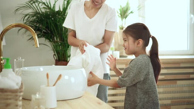 Washing hands daily routine. Spbd Joyful Asian girl with long ponytail wipes clean palms with tidy towel dancing with mother in contemporary bathroom