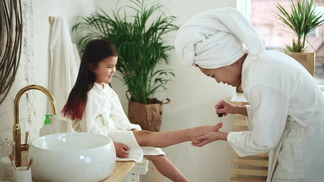 Pedicure mother daughter at home. Spbd Careful Asian woman colors nails on toes of little girl with nail-polish playing beauty salon in light spacious bathroom