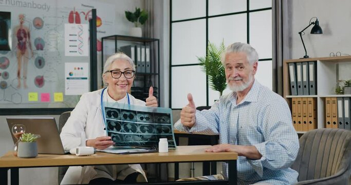 Beautiful smiling experienced respected woman-doctor in glasses with x-ray scan posing on camera together with positive mature bearded man and showing thumbs up