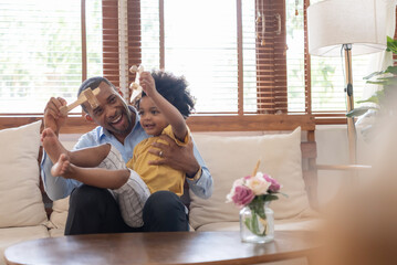 Cheerful African American Father and little son sitting on sofa playing wooden airplane toy at home...