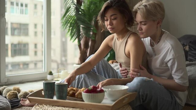 Two lesbian women kissing and eating croissants while having breakfast in bed at home spbd. Closeup view of young attractive females kiss and eat delicious food, look with smiles and sit in light room