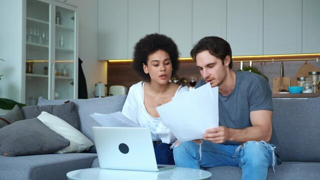 Happy multiethnic couple sitting on sofa in living room holding paper sheets of documents and comparing content on monitor screen while taking notes. Beautiful young couple comparing utility bills