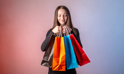 Young pretty woman in a black dress is holding a colorful paper bags with purchases from a shop in her hands on a blue pink background. Concept of Black Friday, sale and shopping