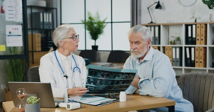Attractive confident respected skilled grey-haired woman-doctor explaining results of x-ray scan to serious mature bearded patient in modern medical office,front view