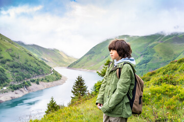 Portrait of a young boy with backpack in a green jacket standing in the mountains near a beautiful lake in French Alps, looking away with a serious face. Hiking with children.