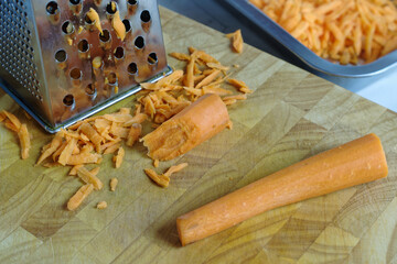Grated carrots in front of a square grater on a wooden cutting board. 