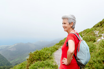 older woman hiker with backpack and gray hair contemplates the mountain landscape on a cloudy day.