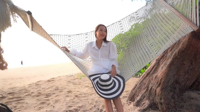A young pretty woman rocks gently in a rope hammock slung over a sandy Southeast Asian beach.