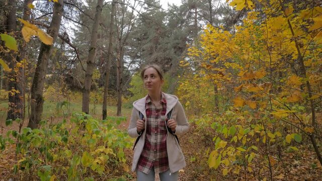Front view of young positive woman with backpack walking in autumn park, forest - wide angle steadicam shot. Active outdoor lifestyle, leisure time, freedom and adventure concept