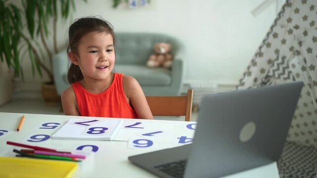 Girl learning math and sitting at table with laptop during online conference in home room spbd. Closeup view of Korean schoolgirl looks at computer screen with smile and shows number, does practice