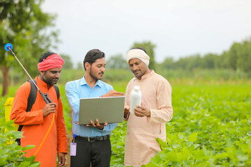 Young indian agronomist giving liquid fertilizer bottle to farmer and showing product information in laptop at green agriculture field.