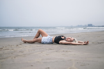 Beautiful slim caucasian young woman with blond hair, wearing denim shorts and black short top. Laying on the sand on the beach and enjoying. Model 
