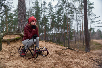 Fototapeta na wymiar A girl in a red jacket and a hat sits on a sled at the forest mountain and waits for the snow