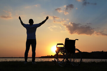 Obraz premium Man raising hands up to sky near wheelchair at sunset, back view. Healing miracle