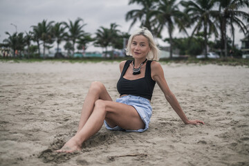 Fototapeta na wymiar Portrait of beautiful fit caucasian young woman with blond hair, wearing denim shorts and short top. Sitting on the beach. Looks directly at camera. 