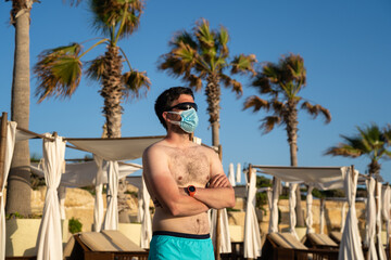 Fototapeta na wymiar Man in medical mask stands alone on empty beach near sun loungers and palm trees on Cyprus, Paphos city during coronavirus quarantine and lockdown. New normal in vacation time after COVID-19 pandemic