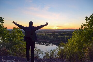 Fototapeta premium Silhouette of a man with a backpack and arms outstretched against the background of a river and an autumn sunset